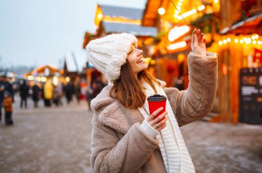 Noel pazarında yürürken sıcak kahve içen bayram ışıklarıyla süslenmiş bir kadın. Yeni yıl. Kış tatili.