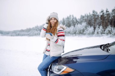 Mutlu kadın bir termos tutuyor ve karlı bir yolda bir arabanın yanında çay içiyor. Dinlenme, rahatlama, seyahat, yaşam tarzı konsepti.