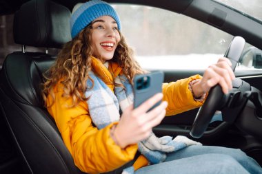 Stylish woman sitting in car dressed in winter clothes using smartphone. Business woman busy driving. Leisure, travel, technology, navigation. clipart