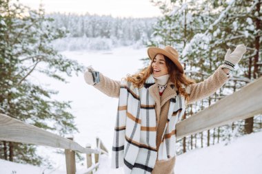 Genç ve güzel bir kadın, kışın kar altında fotoğraf çektirirken gülümser. Orman kızı beyaz parkta yürüyor. Boho tarzı.