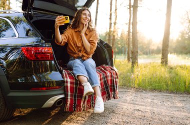 Mutlu Kadın arabanın bagajında dinleniyor ve selfie çekiyor. Selfie zamanı. Yaşam tarzı, seyahat, turizm, doğa, aktif yaşam.