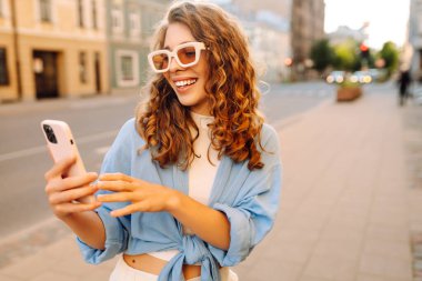 Sokakta elinde telefonla bekleyen güzel bir genç kadının fotoğrafı. Teknoloji kavramı kullan.
