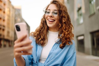 Sokakta elinde telefonla bekleyen güzel bir genç kadının fotoğrafı. Teknoloji kavramı kullan.