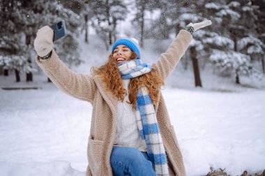 Kış karlı ormanlarda akıllı telefondan selfie çeken gülümseyen genç bir kadın. Kış tatili, tatil, turizm konsepti.