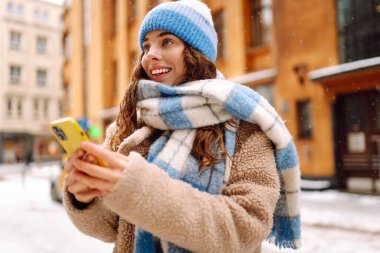 Genç bir kadın şehirde yürüyor ve akıllı telefondan sosyal ağları izliyor, internette sohbet ediyor ve gülümsüyor. Kış havası. Yaşam tarzı, seyahat, turizm, teknoloji