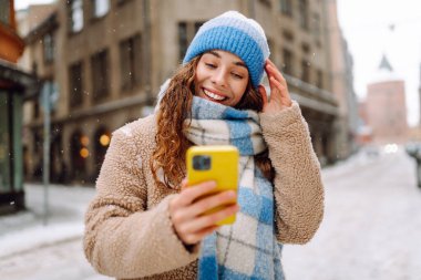 Genç bir kadın şehirde yürüyor ve akıllı telefondan sosyal ağları izliyor, internette sohbet ediyor ve gülümsüyor. Kış havası. Yaşam tarzı, seyahat, turizm, teknoloji