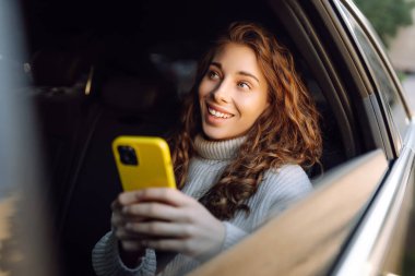Bir arabanın arka koltuğunda elinde akıllı telefonu olan genç bir kadın. Arabayla yolculuk. İletişim çevrimiçi.