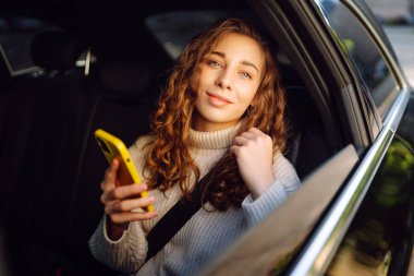 Bir arabanın arka koltuğunda elinde akıllı telefonu olan genç bir kadın. Arabayla yolculuk. İletişim çevrimiçi.