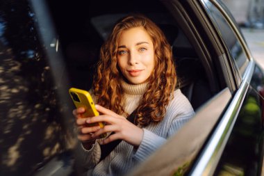 Bir arabanın arka koltuğunda elinde akıllı telefonu olan genç bir kadın. Arabayla yolculuk. İletişim çevrimiçi.