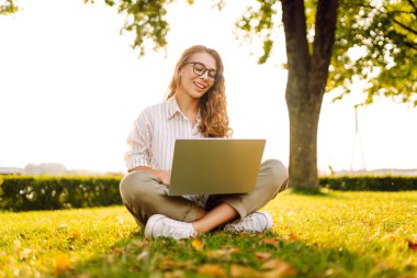 Yeşil bir açıklıkta dizüstü bilgisayarla oturan mutlu kadın. Serbest çalışan, online eğitim. İletişim çevrimiçi.