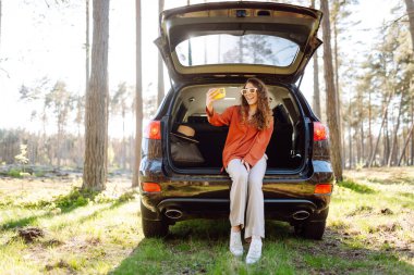 Mutlu kadın arabasının bagajında oturup selfie çekiyor. Arabayla seyahat ederken selfie. Seyahat, hafta sonu.