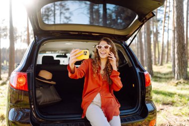 Mutlu kadın arabasının bagajında oturup selfie çekiyor. Arabayla seyahat ederken selfie. Seyahat, hafta sonu.
