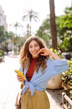 Yaşlı caddede akıllı telefon kullanarak yürüyen gülümseyen genç bir kadın. İletişim, sosyal ağlar, online alışveriş kavramı.