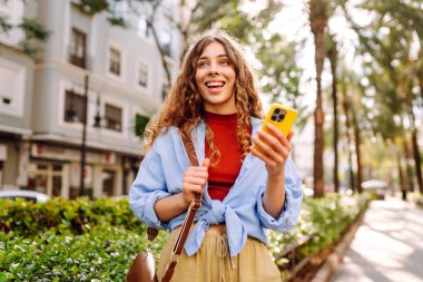 Yaşlı caddede akıllı telefon kullanarak yürüyen gülümseyen genç bir kadın. İletişim, sosyal ağlar, online alışveriş kavramı.