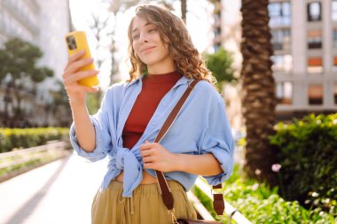 Yaşlı caddede akıllı telefon kullanarak yürüyen gülümseyen genç bir kadın. İletişim, sosyal ağlar, online alışveriş kavramı.