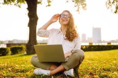 Genç kadın çimlerin üzerinde oturuyor, dizüstü bilgisayarla çalışıyor. Çevrimiçi eğitim, Serbest çalışma, teknoloji kavramı.