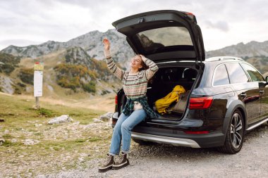 Mutlu kadın arabasının bagajında oturuyor, kayalık araziden geçen virajlı bir yolun kenarına park etmiş. Yaşam tarzı, seyahat, turizm, doğa, aktif yaşam