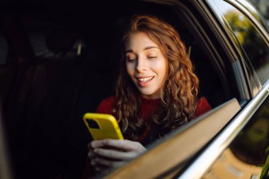 Bir arabanın arka koltuğunda oturan ve kısa mesaj okuyan genç bir kadın. Teknoloji, blog yazma, iş konsepti.