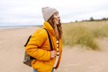 Soğuk ve bulutlu bir günde sırt çantasıyla sahilde yürüyüş yapan çekici, sağlıklı bir kadın. Yaşam tarzı, seyahat, turizm, doğa, aktif yaşam.