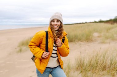 Soğuk ve bulutlu bir günde sırt çantasıyla sahilde yürüyüş yapan çekici, sağlıklı bir kadın. Yaşam tarzı, seyahat, turizm, doğa, aktif yaşam.