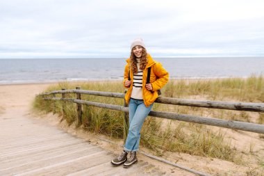 Soğuk ve bulutlu bir günde sırt çantasıyla sahilde yürüyüş yapan çekici, sağlıklı bir kadın. Yaşam tarzı, seyahat, turizm, doğa, aktif yaşam.