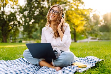 Genç bayan kulaklıklar oturup müzik dinlerler cep telefonu kullanırlar. Dinle, müzik parkta yeşil çimlerin üzerinde çalışıyor, çalışıyor. Eğitim, iş, blog veya serbest çalışma kavramı.
