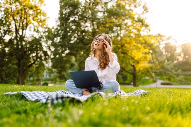 Genç bayan kulaklıklar oturup müzik dinlerler cep telefonu kullanırlar. Dinle, müzik parkta yeşil çimlerin üzerinde çalışıyor, çalışıyor. Eğitim, iş, blog veya serbest çalışma kavramı.