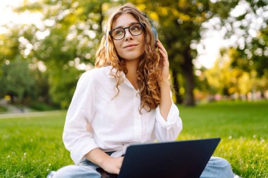 Genç bayan kulaklıklar oturup müzik dinlerler cep telefonu kullanırlar. Dinle, müzik parkta yeşil çimlerin üzerinde çalışıyor, çalışıyor. Eğitim, iş, blog veya serbest çalışma kavramı.