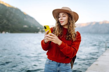 Sırt çantalı ve sıcacık giysili genç bayan gezgin göle karşı iskelede duruyor ve telefon kullanıyor. Avrupa seyahati. Yaşam tarzı, tatil, turizm, doğa, aktif hayat. Günlük tutma.