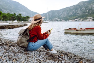Sırt çantalı ve sıcacık giysili genç bayan gezgin göle karşı iskelede duruyor ve telefon kullanıyor. Avrupa seyahati. Yaşam tarzı, tatil, turizm, doğa, aktif hayat. Günlük tutma.