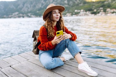 Sırt çantalı ve sıcacık giysili genç bayan gezgin göle karşı iskelede duruyor ve telefon kullanıyor. Avrupa seyahati. Yaşam tarzı, tatil, turizm, doğa, aktif hayat. Günlük tutma.