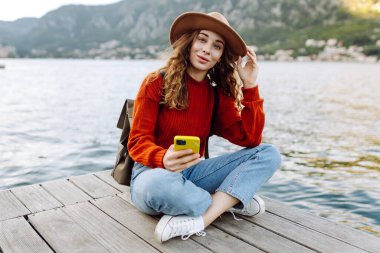 Sırt çantalı ve sıcacık giysili genç bayan gezgin göle karşı iskelede duruyor ve telefon kullanıyor. Avrupa seyahati. Yaşam tarzı, tatil, turizm, doğa, aktif hayat. Günlük tutma.