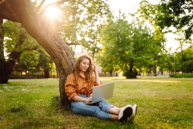 Dışarıda dizüstü bilgisayarı olan yeşil çimlerde oturan güzel kadın. Çevrimiçi eğitim, Serbest çalışma, teknoloji kavramı.