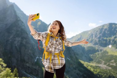 Sarı sırt çantalı ve elinde telefon olan bir kadın yolcu selfie çekiyor. Uçurumun tepesindeki mutlu kadın dağ manzarasının tadını çıkarıyor, ünlü turistik mekanda yürüyüş yapıyor..