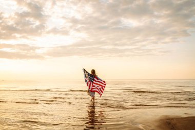 Okyanusta yürüyen Amerikan bayrağı taşıyan genç bir kadın. 4 Temmuz. Amerika Bağımsızlık Günü konsepti.