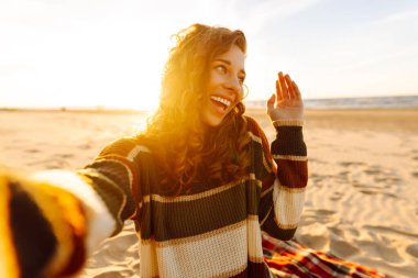 Gülümseyen bir kadın cep telefonundaki kameraya poz verirken sakin okyanuslu kasvetli, soğuk kumlu bir sahilde selfie çekiyor. Gün batımı. Bloglama, seyahat, konsept.