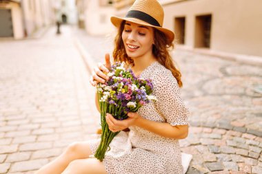 Yazlık kıyafetli güzel bir kadın gülümseyerek sokaklarda çiçeklerle mutlu bir şekilde yürüyor. Moda tarzı.