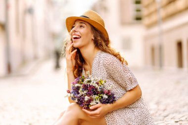 Yazlık kıyafetli güzel bir kadın gülümseyerek sokaklarda çiçeklerle mutlu bir şekilde yürüyor. Moda tarzı.