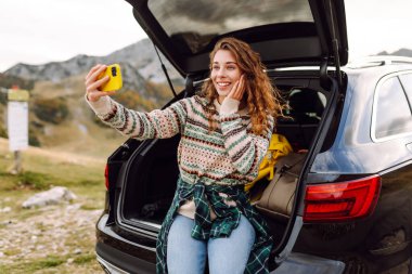 Kafkasyalı genç bir kadın, doğada bir arabanın bagajında otururken akıllı telefon kullanıyor. Seyahat, hafta sonu, doğa, rahatlama ve yaşam tarzı konsepti.