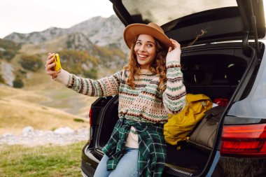 Kafkasyalı genç bir kadın, doğada bir arabanın bagajında otururken akıllı telefon kullanıyor. Seyahat, hafta sonu, doğa, rahatlama ve yaşam tarzı konsepti.