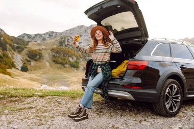 Kafkasyalı genç bir kadın, doğada bir arabanın bagajında otururken akıllı telefon kullanıyor. Seyahat, hafta sonu, doğa, rahatlama ve yaşam tarzı konsepti.