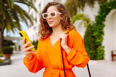 Dışarıda yürürken cep telefonunda gülen ve mesaj atan genç bir kadın. Yaşam tarzı, seyahat, turizm, teknoloji, blog yazma, iletişim kavramı.