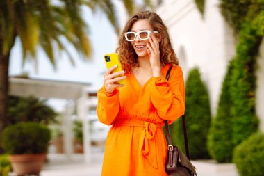 Dışarıda yürürken cep telefonunda gülen ve mesaj atan genç bir kadın. Yaşam tarzı, seyahat, turizm, teknoloji, blog yazma, iletişim kavramı.