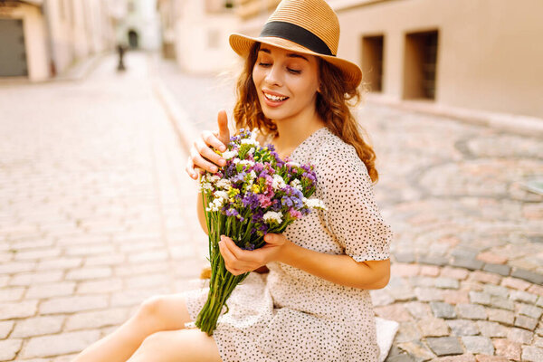 Молодая женщина в летнем наряде счастливо улыбается, гуляя с цветами по городской улице. Стиль моды.