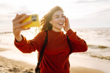 Genç bir kadın gün batımında kumlu bir sahilde neşeli bir selfie çekiyor. Selfie zamanı. Soğuk rüzgarlı hava. Bloglama, teknoloji, yaşam tarzı, seyahat.