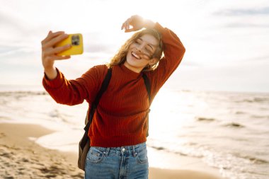 Genç bir kadın gün batımında kumlu bir sahilde neşeli bir selfie çekiyor. Selfie zamanı. Soğuk rüzgarlı hava. Bloglama, teknoloji, yaşam tarzı, seyahat.