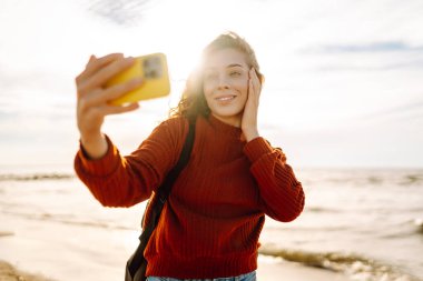 Genç bir kadın gün batımında kumlu bir sahilde neşeli bir selfie çekiyor. Selfie zamanı. Soğuk rüzgarlı hava. Bloglama, teknoloji, yaşam tarzı, seyahat.