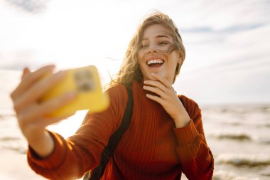 Genç bir kadın gün batımında kumlu bir sahilde neşeli bir selfie çekiyor. Selfie zamanı. Soğuk rüzgarlı hava. Bloglama, teknoloji, yaşam tarzı, seyahat.