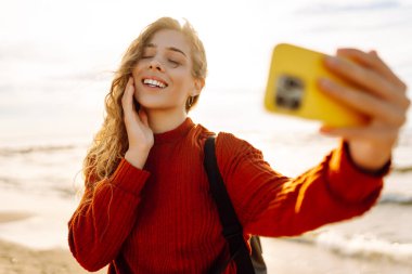 Genç bir kadın gün batımında kumlu bir sahilde neşeli bir selfie çekiyor. Selfie zamanı. Soğuk rüzgarlı hava. Bloglama, teknoloji, yaşam tarzı, seyahat.
