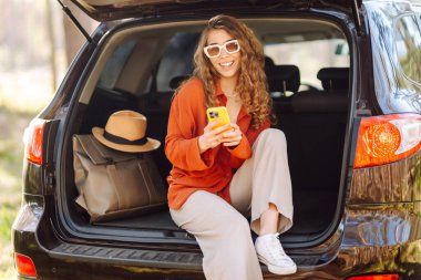 Güzel bir kadın açık havada bir arabanın bagajında oturuyor ve akıllı telefon kullanıyor. Seyahat, hafta sonu, doğa, rahatlama ve yaşam tarzı konsepti. Bloglama, iletişim.